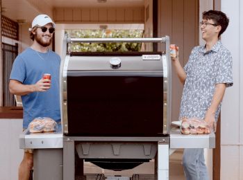 Two people standing by a grill, each holding a drink. They appear to be preparing for a barbecue with burger buns and other items on the grill sides.