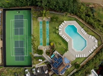 An aerial view of a recreational area with a tennis court, shuffleboard courts, a swimming pool, lounge chairs, and a building with solar panels.