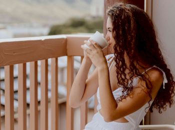 A woman with curly hair in a white dress sits on a balcony, drinking from a mug, with scenic mountains in the background and clear skies.