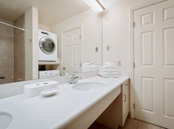 This image features a clean bathroom with a double sink, a stackable washer and dryer, white towels, and a large mirror above the countertop.