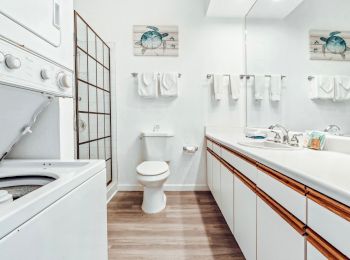 This is a bright, clean bathroom with a washer and dryer, a sink, a toilet, and a shower. Linen towels and decorative artwork are visible.