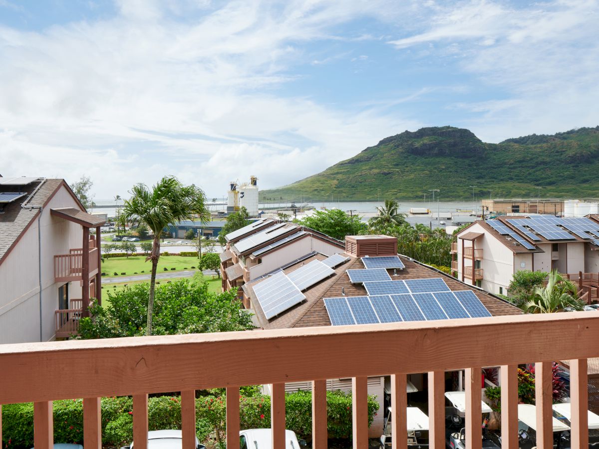 A scenic view of buildings with solar panels, lush greenery, mountains in the background, and a vivid sky, as seen from a wooden balcony.
