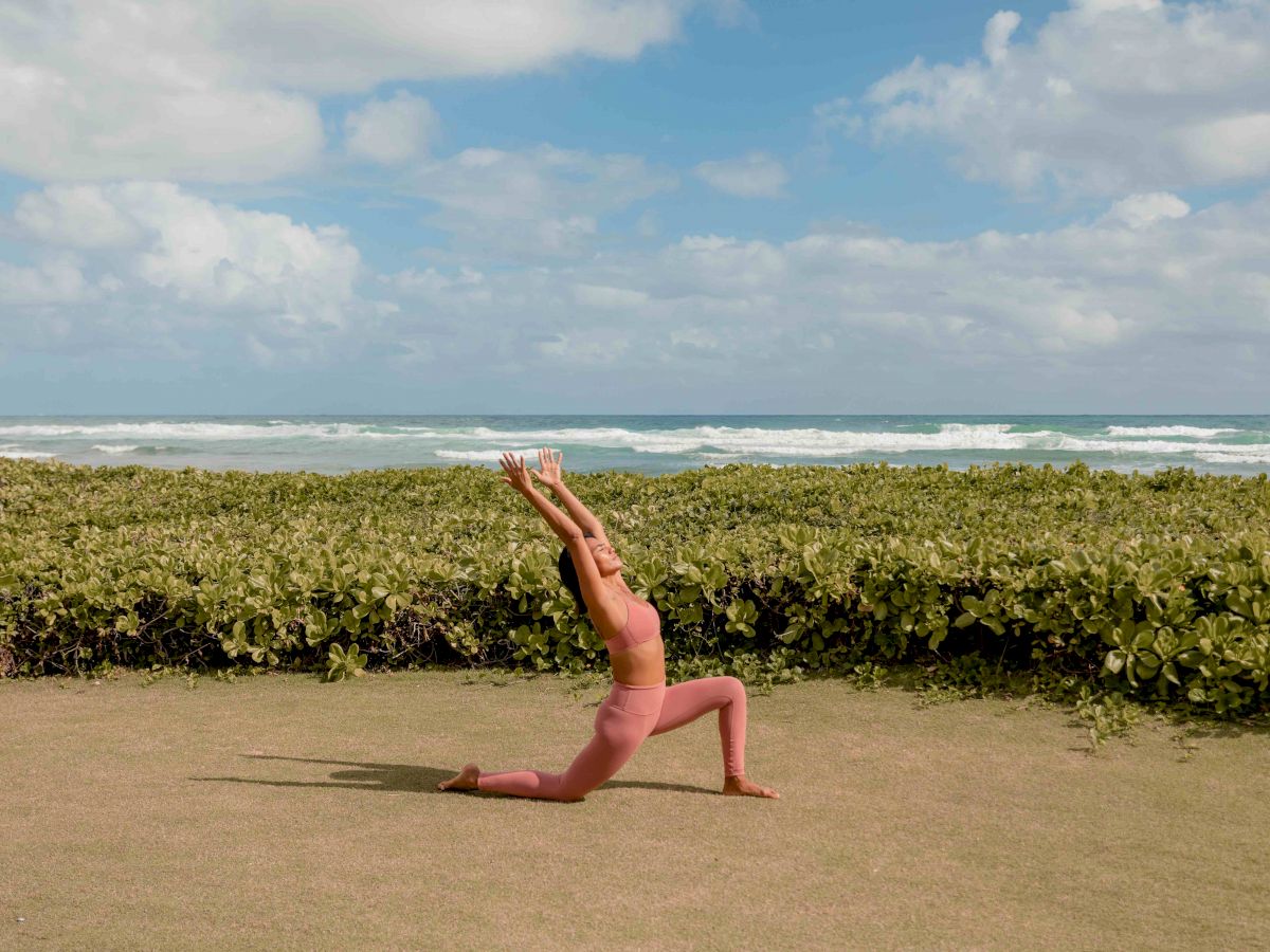 A person in pink athletic attire is practicing a yoga pose on a grassy area near the beach, with the ocean and sky in the background.