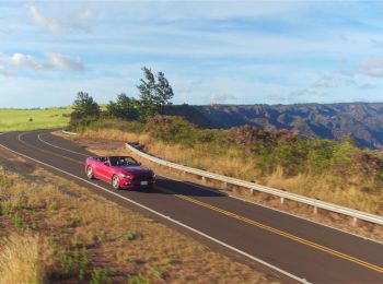 A red convertible car is driving on a winding road through a scenic landscape with mountains and open fields under a blue sky with scattered clouds.