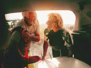 An elderly couple is seen through the back of a car, smiling and preparing for a day with a surfboard and gear. The setting is outdoor adventure.