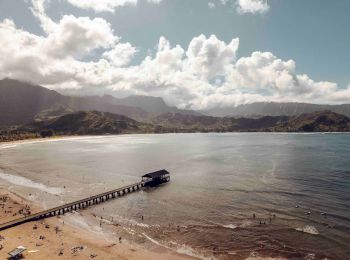 A scenic beach with a wooden pier extending into calm water, people on the beach, mountains in the background, and fluffy clouds in the sky, always ending the sentence.