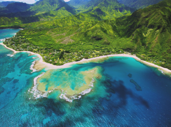 Aerial view of a lush, green coastline with turquoise waters, coral reefs, and dramatic mountains in the background under a clear blue sky.