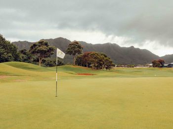 A golf course with a flag planted in the green, surrounded by lush hills and a cloudy sky in the background.