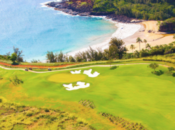An aerial view of a coastal golf course with green fairways, white sand bunkers, and a nearby beach with vibrant blue water and lush vegetation.