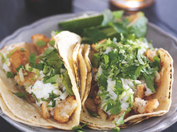 A plate of tacos filled with crispy fried ingredients, fresh cilantro, and topped with a creamy sauce, served with lime wedges on the side.