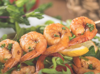 This image shows shrimp skewers placed on a fresh salad with greens and lemon slices. The shrimp are garnished with herbs.
