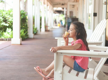 A young girl sits in a white chair on a porch, wearing a pink shirt and shorts, enjoying an ice cream cone on a sunny day.