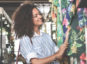 A woman in a blue striped shirt is smiling and browsing colorful floral fabrics at what looks like a market or store.