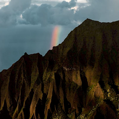 The image shows a rugged mountain landscape with sharp ridges, lit dramatically, and a rainbow appearing in the background, against a cloudy sky.