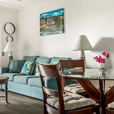 The image features a cozy living-dining area with a light blue sofa, a glass-top dining table, wooden chairs, and a vibrant painting on the wall.
