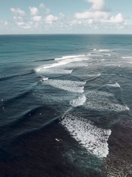An aerial view of the ocean showing waves breaking towards the shore with surfers catching the waves under a partly cloudy sky.