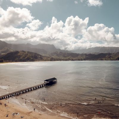 A scenic beach with a pier extending into the water, surrounded by mountains and cloudy skies. People are relaxing on the beach and swimming.
