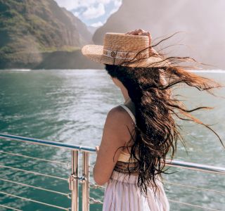 A woman with long, flowing hair and a hat stands on a boat, gazing at the scenic mountainous landscape and water under a partly cloudy sky.
