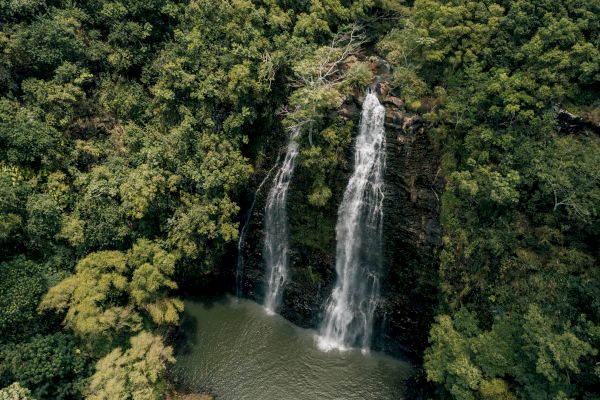 Aerial view of a waterfall cascading into a small pool, surrounded by dense, green forest. The waterfall splits into two separate streams as it descends.
