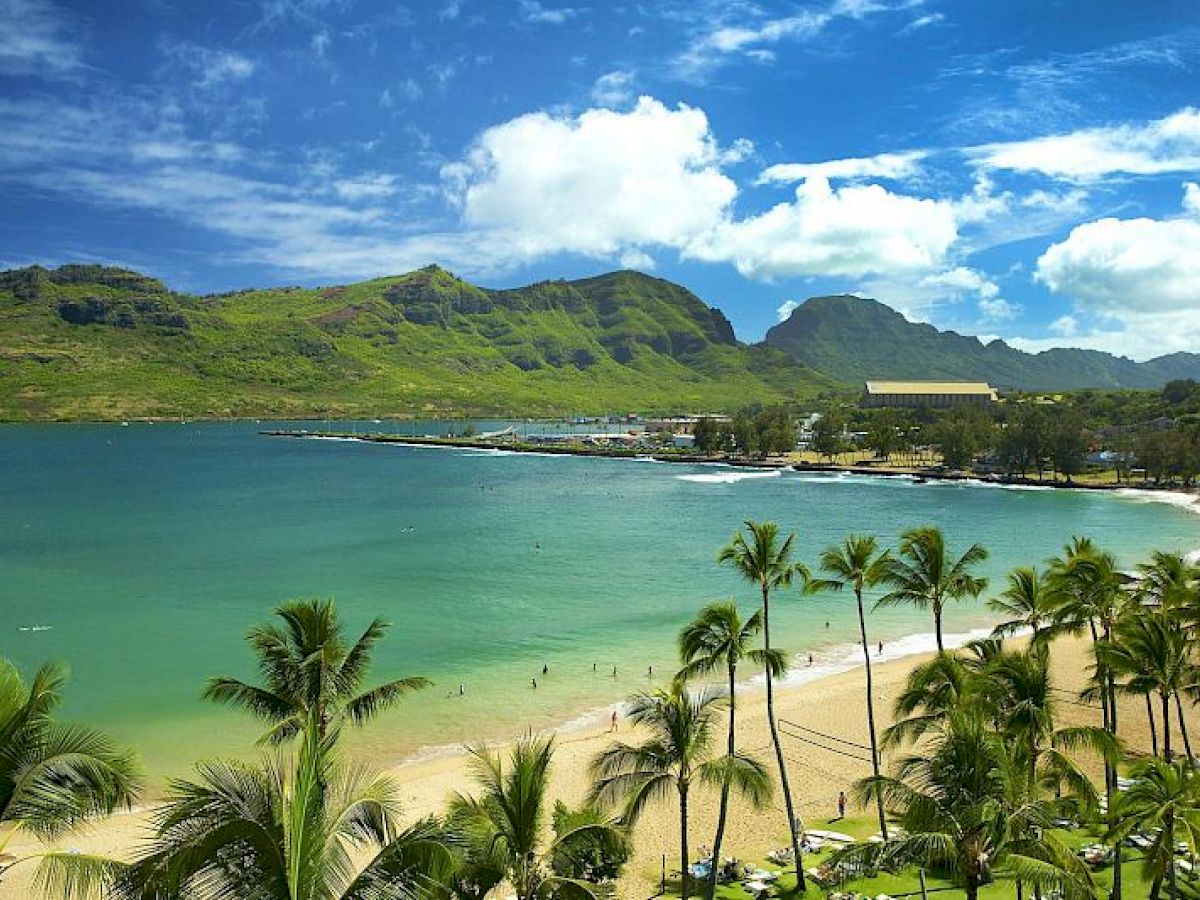 This image shows a beautiful beach with golden sand, palm trees, and clear blue waters, with green mountains and a partly cloudy sky in the background.