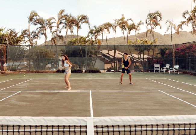 Two people are playing tennis on an outdoor court, with palm trees and mountains in the background, under a clear sky.