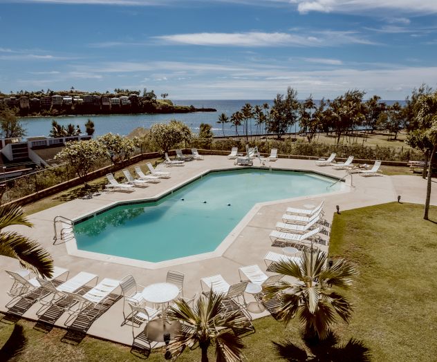 An outdoor swimming pool surrounded by sun loungers, palm trees, and a few tables, with a scenic coastline and ocean in the background.
