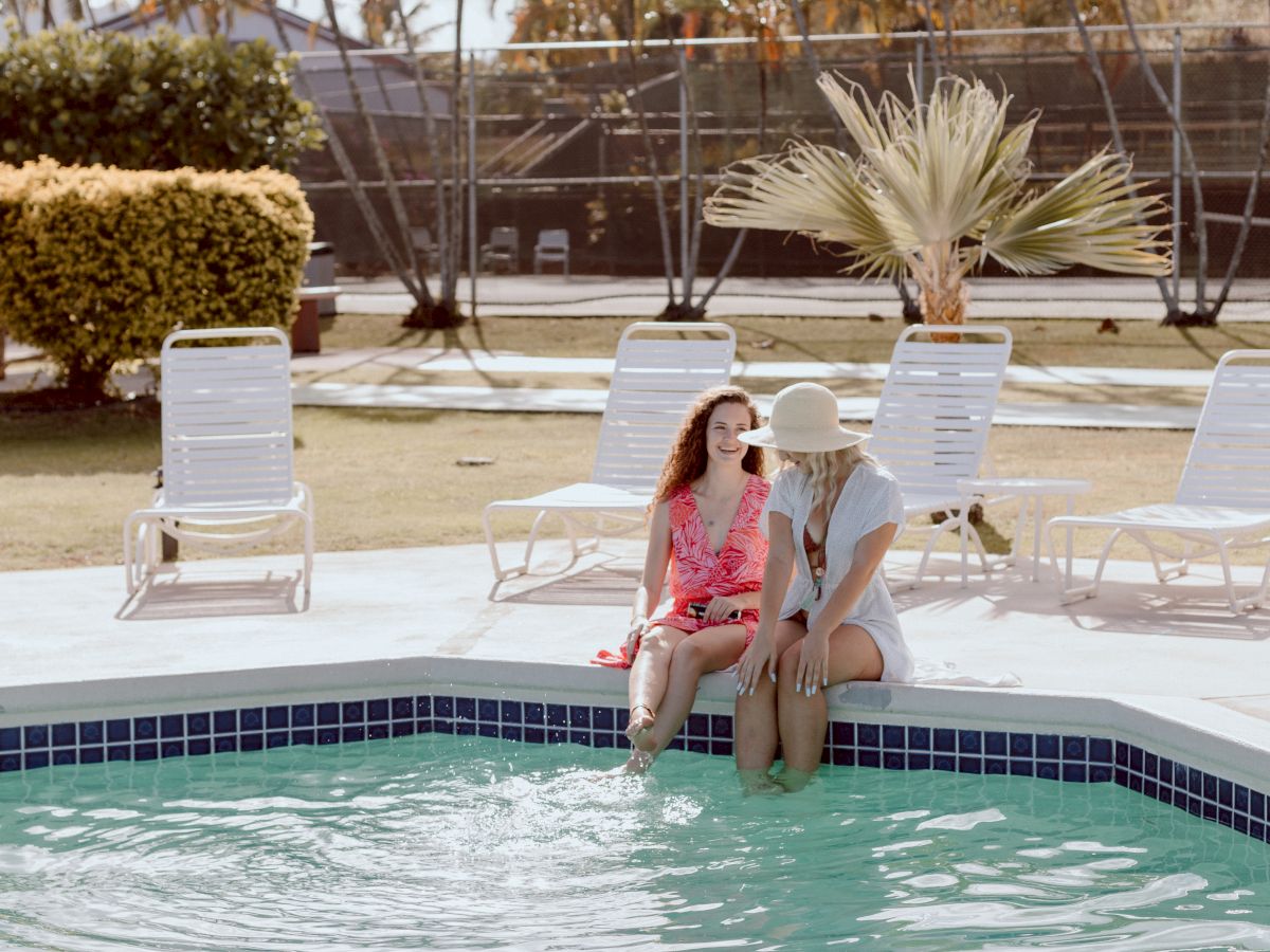 Two individuals sit at the edge of a pool surrounded by lounge chairs, with palm trees in the background.